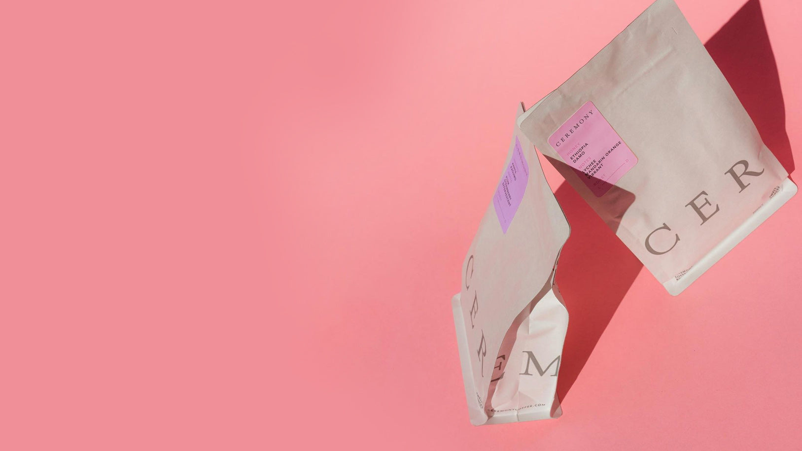 Two beige coffee bags with purple labels reading CEREMONY on a pink background with shadows cast to the right.