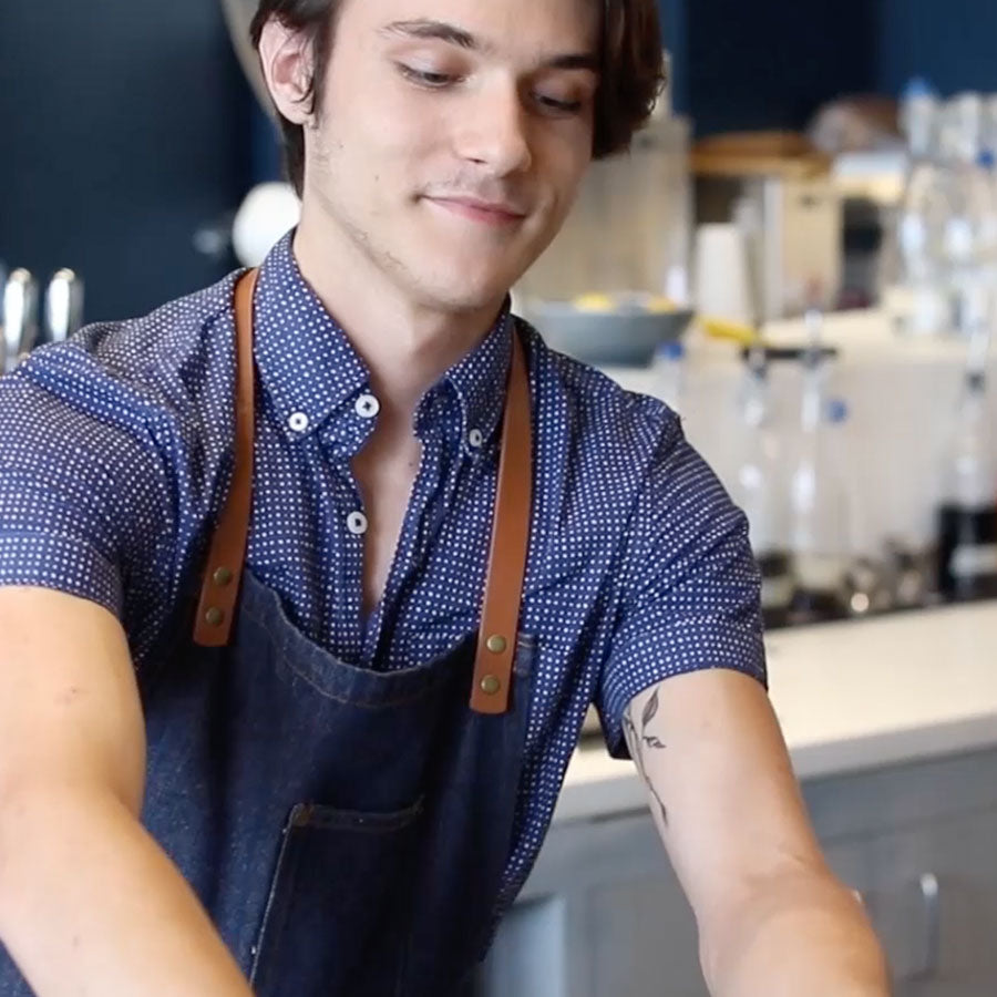 A young man in a blue polka dot shirt and denim apron works at a coffee shop counter, smiling and focused on his task.